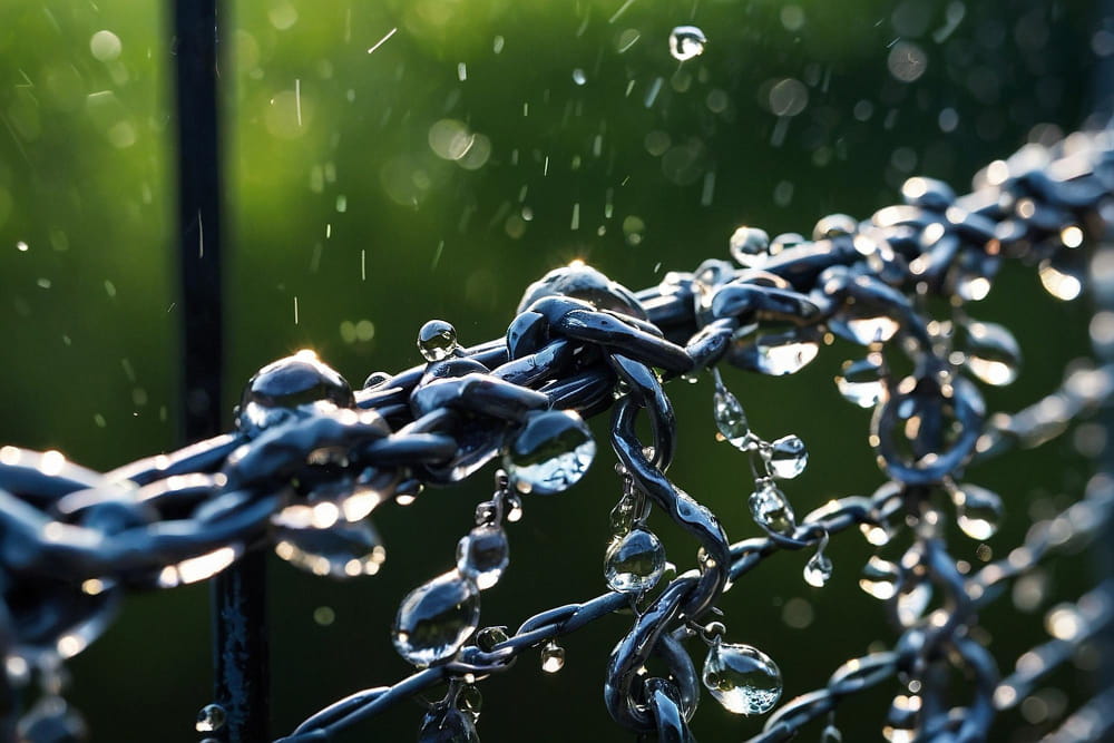 Macro view of sparkling water droplets clinging to dark metallic rain chains during a sunlit shower