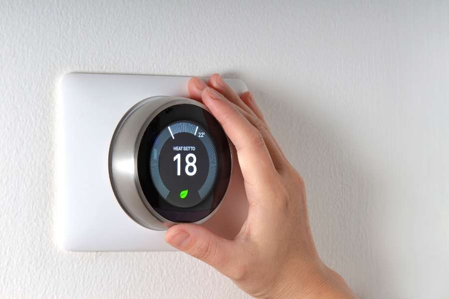 A close-up view of a hand adjusting the temperature on a modern, round smart thermostat mounted on a white wall.