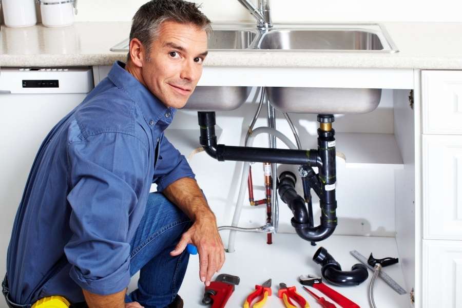 A smiling plumber kneeling next to tools while handling a major plumbing emergency under a sink.